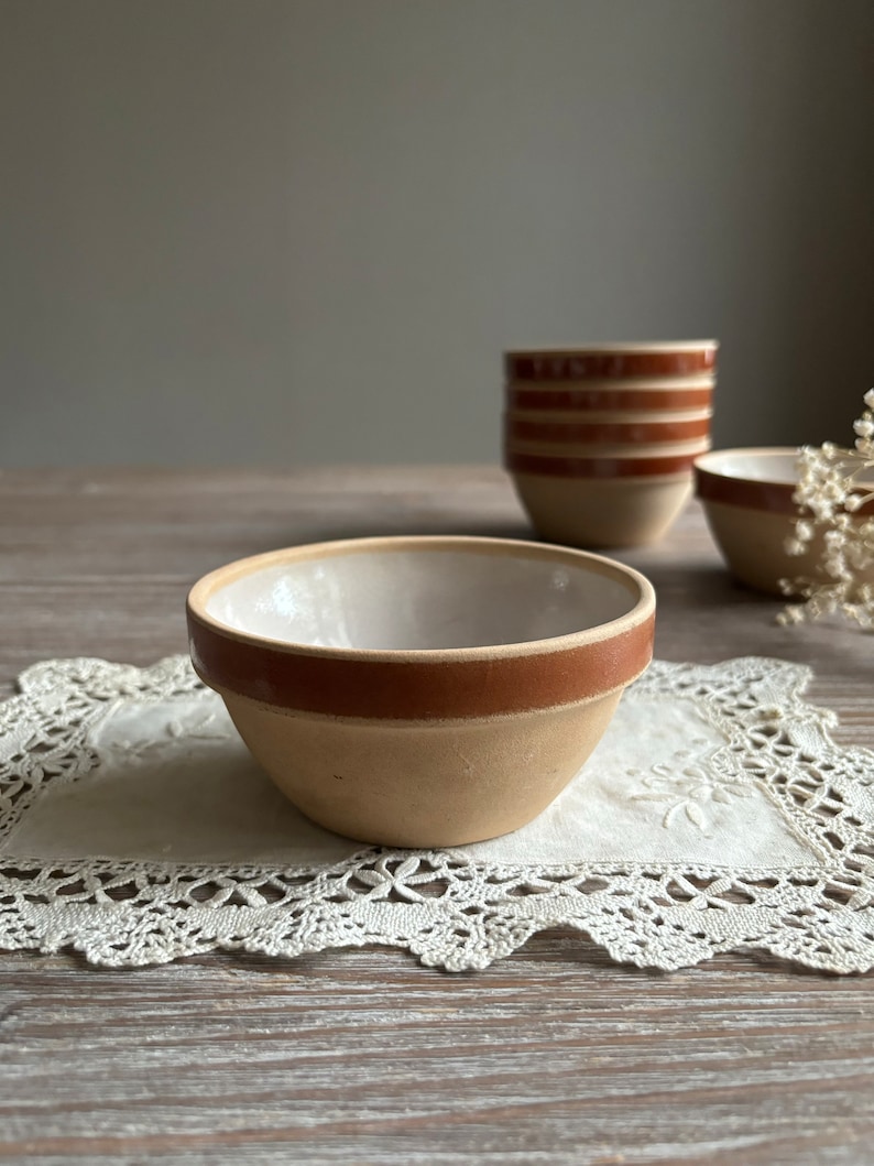 May include: A set of small, white ceramic bowls with a brown rim. The bowls are sitting on a white lace doily on a wooden table.