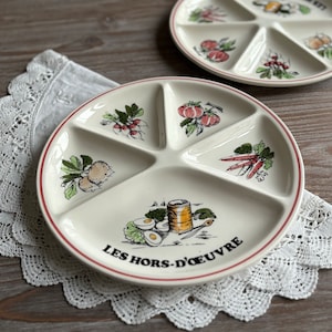 May include: A white ceramic divided serving plate with a red rim. The plate has five sections and features a colourful illustration of vegetables and the text "LES HORS-D'ŒUVRE".