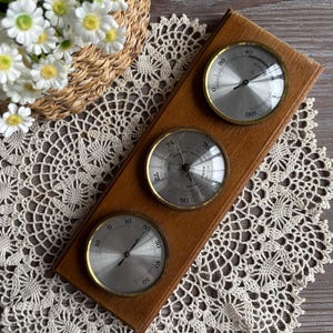Puede incluir: Estación meteorológica de madera con tres medidores circulares. Los medidores tienen caras plateadas y bordes dorados. El medidor superior muestra lecturas de 0 a 100, el del medio de 72 a 80 y el inferior de 0 a 50. Sobre un mantel de encaje.