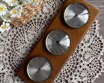 May include: A wooden weather station with three circular gauges. The gauges have silver faces and gold-colored rims. The top gauge reads from 0 to 100, the middle gauge reads from 72 to 80, and the bottom gauge reads from 0 to 50. The weather station is on a lace doily.