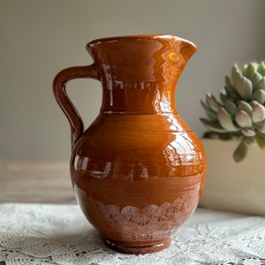 May include: A glazed, brown ceramic pitcher with a handle. The pitcher has a wide base that tapers to a narrow neck and flared rim. The surface reflects light, suggesting a glossy finish. The pitcher is likely for decorative or functional use.