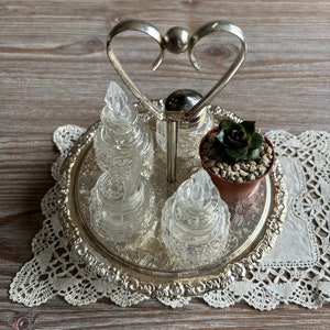 May include: A silver heart-shaped condiment holder with four glass cruets and a small potted succulent plant. The holder is on a silver tray with a decorative edge.