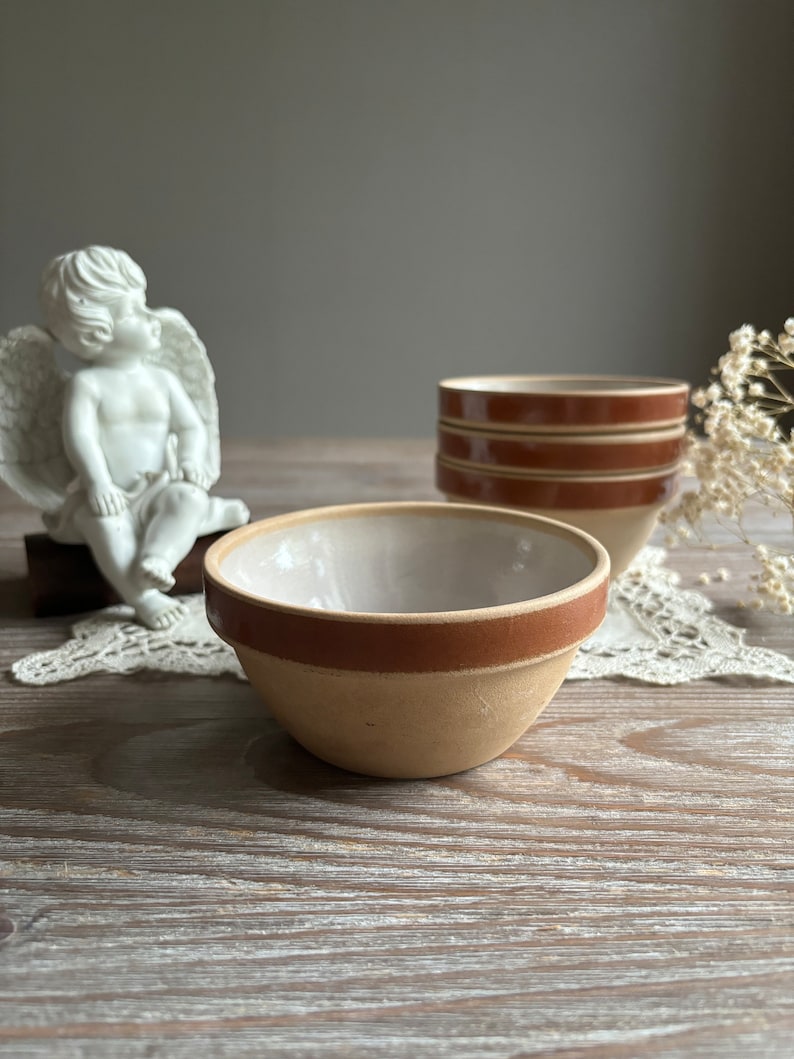 May include: Three brown and white ceramic bowls with a brown rim. The bowls are stacked on top of each other. The bowls are on a wooden table with a white lace doily.