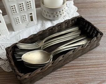 May include: A brown wicker basket filled with silver spoons and forks. The basket is sitting on a wooden table.