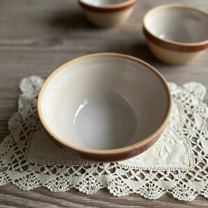 May include: Three white ceramic bowls with brown rims. The bowls are on a white lace doily on a wooden surface.