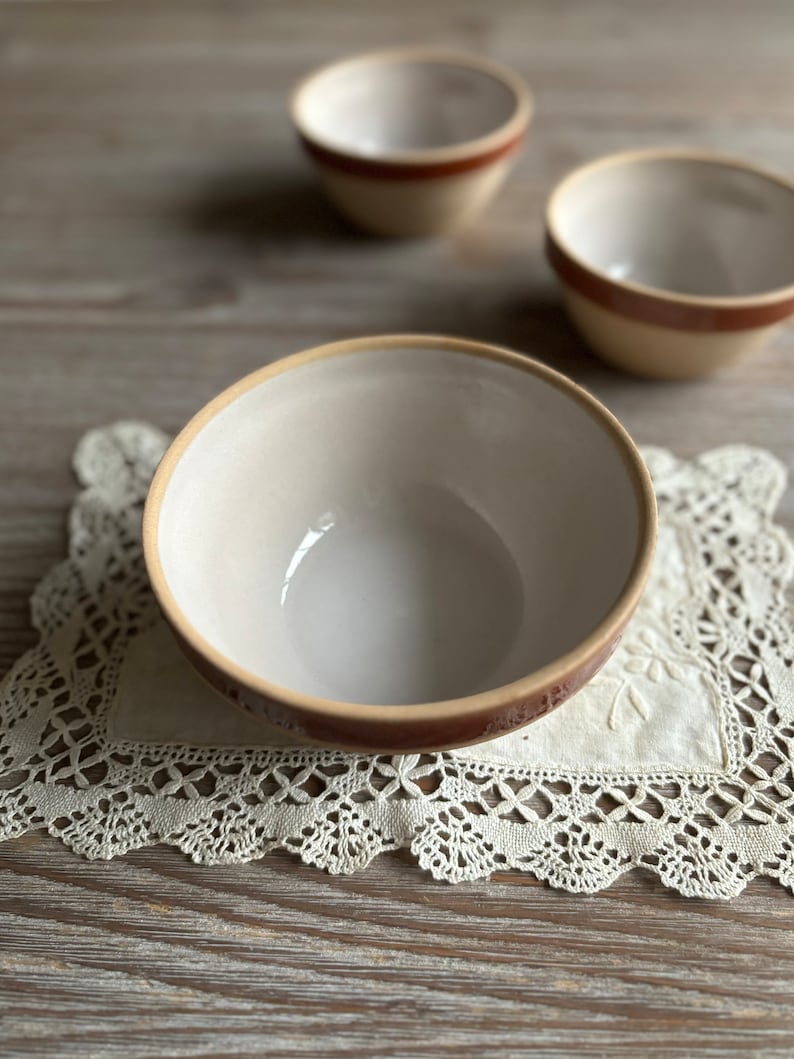 May include: Three white ceramic bowls with brown rims. The bowls are on a white lace doily on a wooden surface.