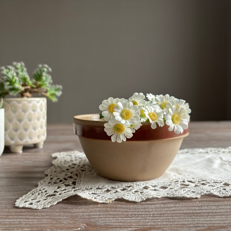May include: A brown and beige ceramic bowl with a brown rim, filled with white daisies. The bowl is sitting on a white crocheted doily on a wooden surface.
