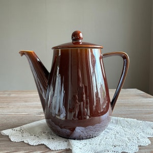 Peut inclure: Une théière en céramique brune avec un corps arrondi et un couvercle avec un bouton. La théière a un bec verseur et une poignée.
