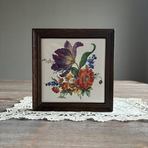 May include: A framed tile featuring a floral design with a purple tulip, red poppy, and other colourful flowers. The tile is set in a dark wood frame.