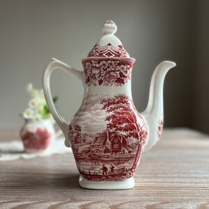 Puede incluir: Una tetera de cerámica blanca con un diseño floral rojo y blanco. La tetera tiene una tapa y un pico. El diseño presenta una cabaña y árboles en un entorno paisajístico.