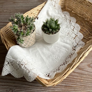 May include: Two small succulent plants in white and patterned pots, sitting on a white doily in a wicker basket.