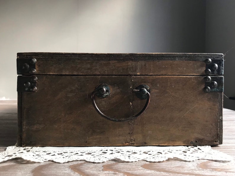 Puede incluir: Una caja de madera con una manija de metal y detalles met&aacute;licos. La caja es marr&oacute;n y tiene un aspecto desgastado.