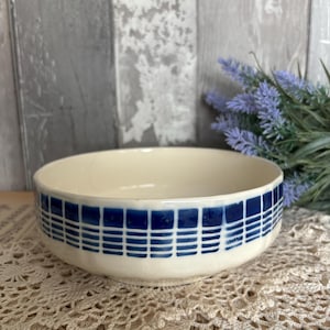 May include: Cream-colored ceramic bowl with a blue grid pattern around the rim. The bowl is set on a cream-colored lace doily. The background features a weathered wooden surface and sprigs of lavender.