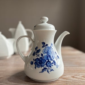 Puede incluir: Una tetera de porcelana blanca con un diseño floral azul. La tetera tiene una tapa y un pico. El diseño floral presenta rosas azules y hojas.