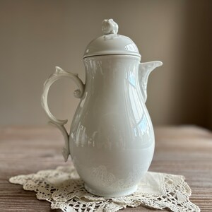 Puede incluir: Cafetera de porcelana blanca con un diseño floral en la tapa y un asa delicada. La cafetera está sobre un encaje blanco.