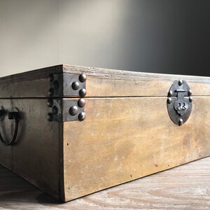 Puede incluir: Una caja de madera con tapa abatible y cierre de metal. La caja tiene refuerzos met&aacute;licos en las esquinas y una manija de metal en el lateral. La caja est&aacute; hecha de madera marr&oacute;n claro y tiene un aspecto r&uacute;stico.
