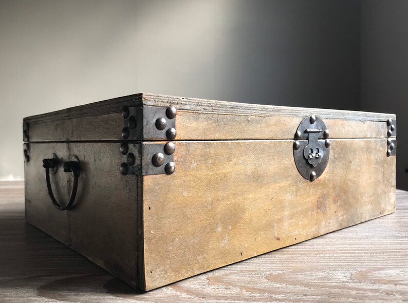 Puede incluir: Una caja de madera con tapa abatible y cierre de metal. La caja tiene refuerzos met&aacute;licos en las esquinas y una manija de metal en el lateral. La caja est&aacute; hecha de madera marr&oacute;n claro y tiene un aspecto r&uacute;stico.