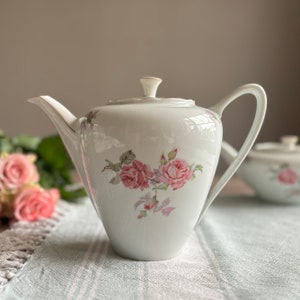 May include: White porcelain teapot with a floral design featuring pink roses and green leaves. The teapot has a lid and a handle.