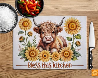 Könnte beinhalten: Rechteckiges Schneidebrett mit einer Aquarellillustration einer Hochlandkuh, umgeben von Sonnenblumen. Der Text "Bless this kitchen" ist in Braun gedruckt. Ein schwarzes und silbernes Messer und Schüsseln mit Essen sind ebenfalls zu sehen.