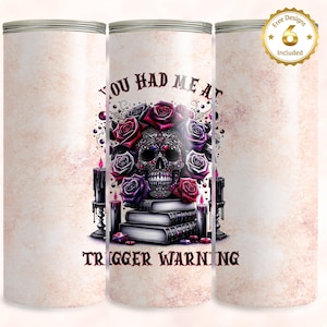 Puede incluir: Un vaso blanco con un diseño de calavera negro y morado con flores y velas. El texto "You had me at trigger warning" está impreso en el vaso.