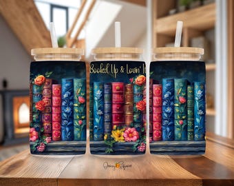 Könnte beinhalten: Drei transparente Glasbecher mit Bambusdeckeln und Strohhalmen. Jeder Becher zeigt eine farbenfrohe Illustration eines Bücherregals voller Bücher und Blumen. Der mittlere Becher trägt den Text "Booked Up & Lovin' It".