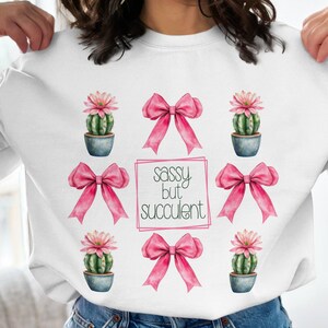 May include: White sweatshirt with a pink bow and cactus design. The text "Sassy but succulent" is printed on the front.