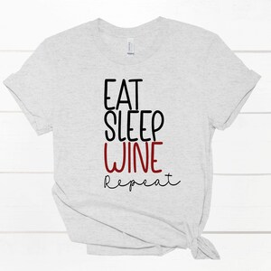 May include: A light grey t-shirt featuring the text "EAT SLEEP WINE REPEAT". The words "EAT" and "SLEEP" are in black, "WINE" is in red, and "repeat" is in black script. The t-shirt is knotted at the bottom.