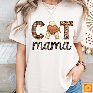 Op de afbeelding: Crèmekleurig T-shirt met de tekst "CAT mama" in bruin script. De letters "CAT" zijn ontworpen met ruit-, stippen- en luipaardprints. Een bruin hart met het woord "cat" staat in het midden.