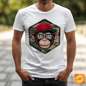 Può includere: T-shirt bianca con un cartone animato di una scimmia con occhiali da sole e un berretto da baseball rosso. La grafica della scimmia è all'interno di un esagono mimetico con le parole "RAD DAD".