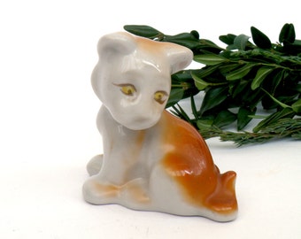 May include: A small, white porcelain figurine of a puppy with brown markings. The puppy is sitting with its head tilted down and has yellow eyes.