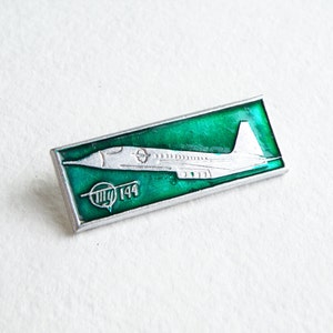 Pode incluir: Pin retangular prateado com fundo verde apresentando um design de avião branco. O pin tem o texto "Tu 144" em branco. Provavelmente um item colecionável vintage.