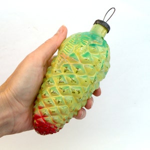 May include: A vintage Christmas ornament shaped like a pinecone. The ornament is painted in shades of yellow and green, with a red base. It has a metal cap and a wire loop for hanging.