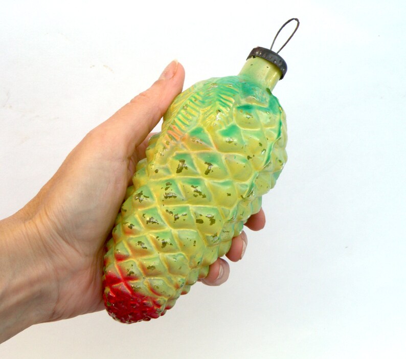 May include: A vintage Christmas ornament shaped like a pinecone. The ornament is painted in shades of yellow and green, with a red base. It has a metal cap and a wire loop for hanging.