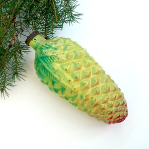 May include: A vintage Christmas ornament shaped like a pine cone. The ornament is primarily yellow with green and red accents, and has a textured surface. A sprig of evergreen needles is in the upper left corner.