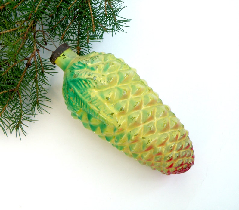 May include: A vintage Christmas ornament shaped like a pine cone. The ornament is primarily yellow with green and red accents, and has a textured surface. A sprig of evergreen needles is in the upper left corner.