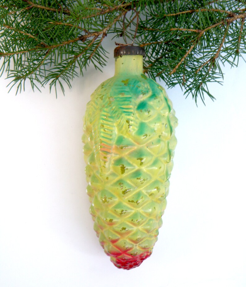 May include: A vintage Christmas ornament shaped like a pine cone, with a textured surface and a gradient of colors. The ornament transitions from yellow-green to red at the base. A sprig of evergreen needles is in the background.