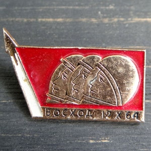 Pode incluir: Um broche de esmalte vermelho e dourado com três astronautas de perfil, com o texto "ВОСХОД 12 X 64" abaixo.