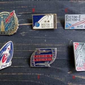 May include: Six vintage enamel pins featuring Soviet space program imagery. The pins depict rockets, spacecraft, and the text "Восток-3", "Восток-4", "Союз-3", "Союз-4", "Союз-5", and "11-12-8-1962".