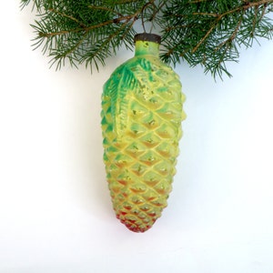 May include: A vintage Christmas ornament shaped like a pineapple. The ornament is yellow with green accents and a red base. It hangs from a green pine branch against a white background. The ornament has a textured surface.
