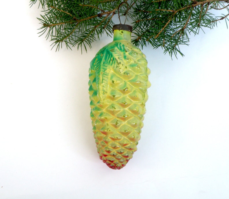 May include: A vintage Christmas ornament shaped like a pineapple. The ornament is yellow with green accents and a red base. It hangs from a green pine branch against a white background. The ornament has a textured surface.