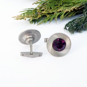 May include: A pair of silver cufflinks with a purple gemstone in the center of one cufflink.