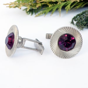 May include: A pair of silver cufflinks with a circular design. Each cufflink features a deep purple rhinestone in the center.