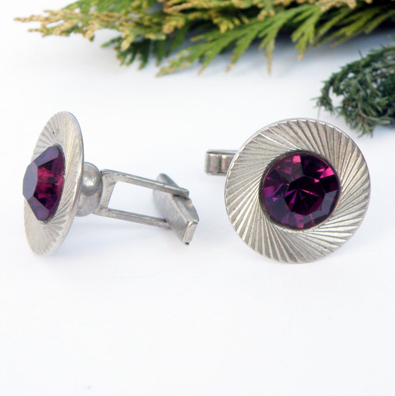 May include: A pair of silver cufflinks with a circular design. Each cufflink features a deep purple rhinestone in the center.