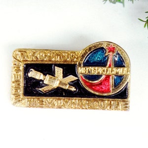 May include: A gold-colored pin with a black and white image of a satellite and the text "Космос Международные" in Cyrillic script. The pin also features a red, white, and blue circle with a star and the text "Интеркосмос" in Cyrillic script.