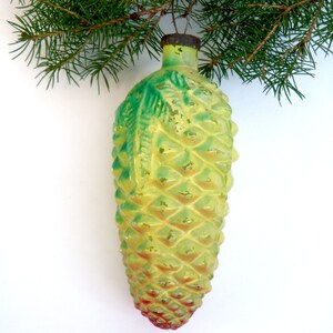 May include: A vintage Christmas ornament shaped like a pinecone, with a textured surface and a mix of yellow, green, and red hues. The ornament is suspended from a metal cap and a green pine branch.