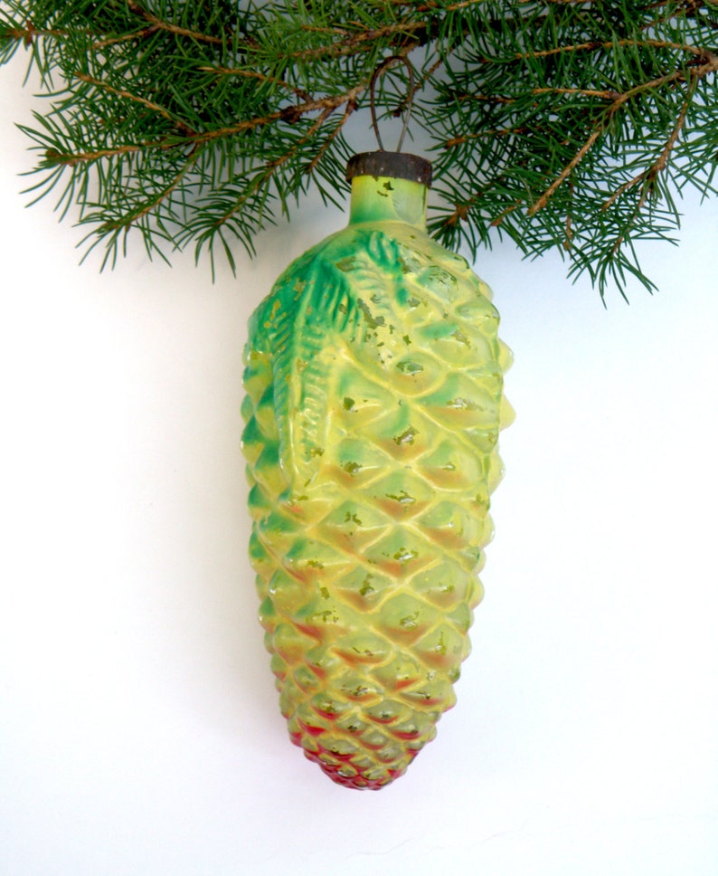 May include: A vintage Christmas ornament shaped like a pinecone, with a textured surface and a mix of yellow, green, and red hues. The ornament is suspended from a metal cap and a green pine branch.