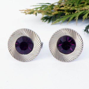 May include: A pair of silver cufflinks with a circular design. Each cufflink features a purple rhinestone in the center.