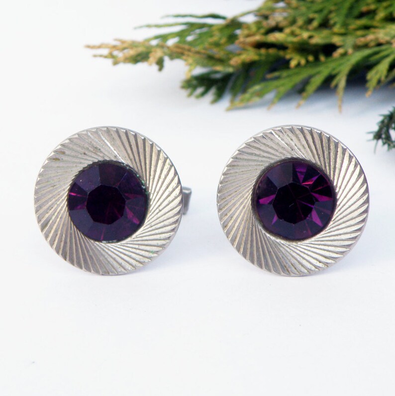 May include: A pair of silver cufflinks with a circular design. Each cufflink features a purple rhinestone in the center.
