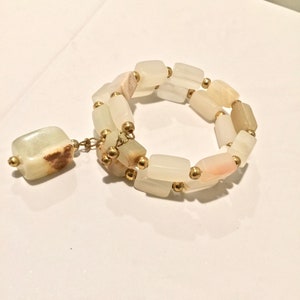 Könnte beinhalten: Ein weißes und hellrosa Stein-Armband mit goldenen Perlen. Das Armband hat einen einzelnen, größeren Stein-Anhänger.