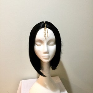 Pode incluir: Uma corrente de cabelo dourada com um design decorativo, usada em uma peruca preta em uma cabeça de manequim branca.